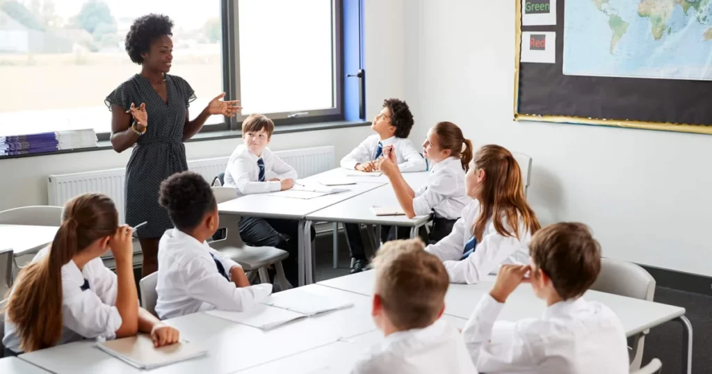 A teacher leading a classroom discussion with pupils in school uniform.